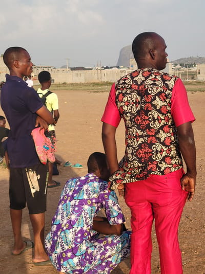 The image depicts several individuals in an open, undeveloped area near what appears to be the outskirts of Ibo, Nigeria. One man, wearing a brightly colored red suit and a floral-patterned shirt, stands with his hand on the shoulder of another individual wearing a patterned purple shirt. Another man is wearing a blue shirt, and the remains of a pair of shoes can be seen. Other individuals are visible in the background. Construction can be seen in the background. The ground is dirt or gravel.