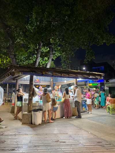 People stand in line at a food stall at night. The stall has a wooden frame and bright lighting. A "Subway" sign is visible further down the street. A large leafy tree is to the left of the stall, and the area is dimly lit beyond the stall. The ground appears to be made of wooden planks. Most of the people in line are facing the stall.