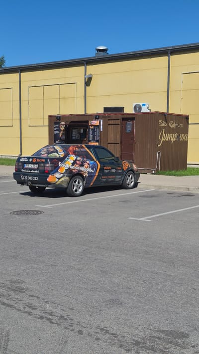 This media file shows a food truck, a dark-colored car with colorful advertisements, parked in an outdoor paved area. The car is decorated with images of sushi and text in Latvian. A brown shipping container, also adorned with text and acting as a food stall, is positioned behind the car. Signs on the container advertise "Suši" (sushi) and provide contact information, including a phone number and a website. The overall setting appears to be a commercial area, with a yellow building visible in the background. The sky is a clear, bright blue, suggesting a sunny day. The image is captured from a slightly elevated perspective, looking down at the scene. The car has a license plate that reads "NY-3159". Text on the car and container includes "Gardi un ar mīlestību!" (Delicious with love!), and lists several locations: Jēkabpils, Līvāni, Madona, Daugavpils. The word "Jumprava" is also visible, possibly indicating a town or origin. The food truck appears to be open for business, with serving windows visible on the container.