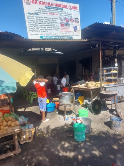 This is an outdoor scene, likely a street market or vendor area in Tanzania, judging by the Swahili text on the prominent sign. The image is oriented sideways. The sun is shining brightly, casting shadows, indicating it's daytime.

The primary subject is a sign for "DR KALULU HERBAL CLINIC," which advertises herbal remedies for various ailments in Swahili. The sign also includes contact information and a Bible verse.

In the foreground, a person in a red shirt and blue shorts is lying down, appearing relaxed or possibly resting. In the background, there are various objects associated with food vending and possibly some form of preparation or storage. These include large metal pots, plastic buckets (some green, one blue), wooden crates, and piles of coconuts and tomatoes. Several other people are visible in the background, some standing and some sitting, suggesting an active market environment. The setting appears somewhat rustic, with a corrugated metal roof visible overhead and a dusty ground surface. The overall mood seems casual and everyday, capturing a snapshot of daily commerce and life.