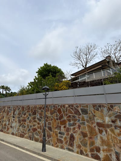 The image depicts a street scene in Sant Andreu de Llavaneres, Spain, featuring a stone wall bordering a road. The wall is constructed of irregular, multi-colored stones and is partially obscured by a grey, metal fence. A streetlight extends from the fence. Beyond the fence and wall are various trees and vegetation. A building is visible in the upper portion of the frame, appearing to be situated on a higher elevation. The sky is overcast with clouds. The image is rotated, with the sky appearing at the bottom and the ground at the top.