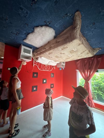 This image depicts a surreal, upside-down bedroom setting in Kamchatka, Russia, where furniture and people are oriented as if gravity were reversed. A large bed, covered with a patterned comforter, is attached to the ceiling. Beside it, a white nightstand with a decorative sphere sits on the "floor" (which is the wall from a normal perspective). A fluffy white rug is placed near the bed.
In the foreground, two individuals are interacting in this inverted environment. One person, wearing a white tank top and denim shorts, reaches towards the nightstand, holding what appears to be a toy. Another person, a child wearing a patterned dress and a pink hat, stands on the wall. Further back, a person wearing a straw hat and a patterned shirt is visible, seemingly observing the scene.
The room has vibrant red walls and a window with red curtains, looking out onto lush green foliage, suggesting it is daytime and the weather is pleasant. The overall impression is one of a playful and disorienting exhibit, likely designed to create a sense of wonder and amusement. There is no visible text in the image.
