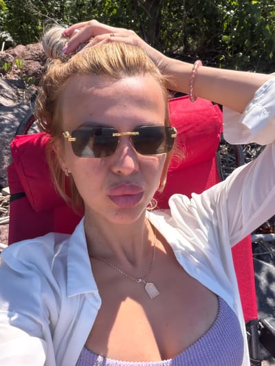 A fair-skinned woman with blonde hair pulled into a messy bun is taking a selfie outdoors. She is wearing large, square-shaped sunglasses with gold trim, a white button-down shirt, and a lavender ribbed bikini top. A gold necklace with a rectangular pendant hangs around her neck. Her left arm is raised behind her head, and her right arm rests on her side. A pink beaded bracelet is visible on her right wrist. She is sitting in a red chair. The background shows green foliage and rocks, suggesting an outdoor, natural setting, possibly a park or a waterfront area.