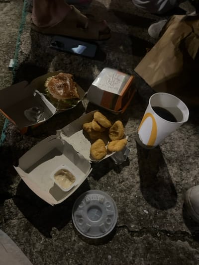 Discarded McDonald's food and containers lie on a rough surface. A burger in its open box, a box of chicken nuggets, a small container of sauce, a drink cup, and a drink lid are scattered around. A brown paper bag is partially visible. A phone and a pair of sandals are nearby. The setting appears to be outdoors, possibly on a sidewalk.