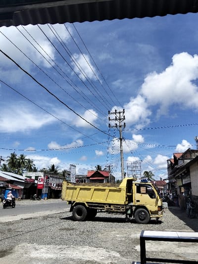 A yellow dump truck sits parked on a gravelly area next to a street with shops and houses. Utility lines crisscross the clear blue sky with fluffy white clouds above the truck. A utility pole is directly behind the vehicle. A person on a scooter rides on the street. A partial view of a corrugated roof overhang is visible at the top of the frame.