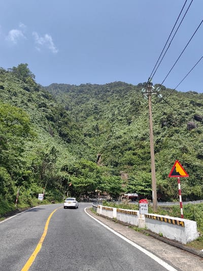 The image depicts a winding mountainous road in Hải Vân Ward, Vietnam, under a clear blue sky with minimal cloud cover. A two-lane asphalt road, marked with a yellow center line and a white shoulder line, curves to the right and recedes into the distance. A white sedan is visible traveling away from the viewer on the right lane of the road.
On the right side of the road, a low concrete barrier with intermittent black and yellow warning stripes lines the edge. Behind this barrier, a white rectangular sign indicates "Km 907" and "HUE 81 km." Further along the right side, a tall wooden utility pole with multiple glass insulators supports three overhead power lines that stretch across the upper portion of the frame. Adjacent to the utility pole, a yellow triangular warning sign with a black border displays an orange and black graphic, indicating a hazard. Below the road level on the right, some small structures or shops with red roofs are partially visible.
The surrounding environment consists of densely vegetated mountainsides, covered in various shades of green foliage and trees, rising steeply on both sides of the road and forming the background. Some exposed rock formations are visible within the heavy vegetation. On the left side of the road, a red and white striped pole is visible, along with other smaller, partially obscured signs.
