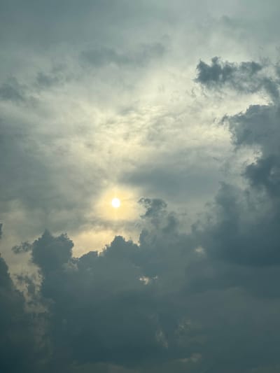 The image shows a sky filled with dramatic clouds. The sun is visible as a bright, almost white circle in the center of the frame, partially obscured by wispy clouds. The surrounding clouds range in color from light gray to dark, ominous gray, suggesting a transition in weather, possibly before or during a storm. The sunlight filters through the clouds, creating a diffused and atmospheric lighting effect. There are no people, objects, or discernible location cues in the image, other than the implied sky. The overall mood is somewhat dramatic and awe-inspiring, with the interplay of light and shadow in the clouds. The time of day is difficult to pinpoint precisely but the quality of light suggests it could be late afternoon or early evening, or a time of day with significant cloud cover. The scene is a natural skyscape. There is no visible text.