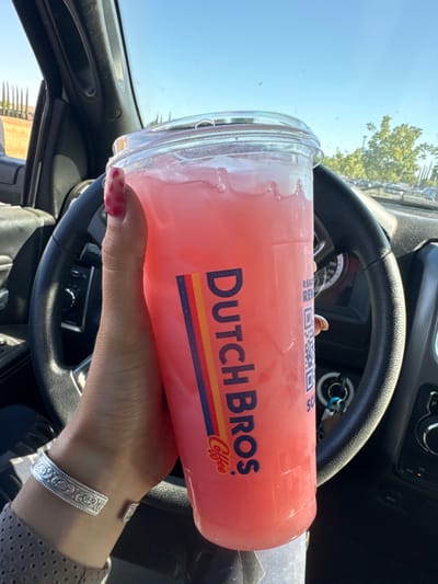 A close-up, eye-level photograph captured from within a vehicle, centered on a human hand holding a clear plastic cup from Dutch Bros Coffee. The cup contains a pink, translucent beverage with ice and displays the brand's distinctive vertical orange, red, and blue striped logo with "DUTCH BROS Coffee" text. A white label with a QR code and partial text "READ REWARDS" is visible on the cup's right side. The hand features a red, mottled-patterned thumbnail and a silver-toned, open-cuff bracelet with an etched design on the wrist. The individual's forearm is covered by a light-colored, textured sleeve.
The vehicle's interior shows a black, multi-spoke steering wheel in the foreground, with portions of the dashboard and controls visible beyond it. A set of keys hangs from the ignition or a key holder to the right of the steering wheel. Through the windshield, a clear blue sky is visible. The external environment on the left consists of a row of slender, dark green coniferous trees on a sunlit, light-colored incline. On the right, green deciduous trees and a paved area consistent with a parking lot or drive-thru lane, with indications of other vehicles, are present. The scene is consistent with a Dutch Bros Coffee location in a commercial area within the city of Rancho Cordova, United States.