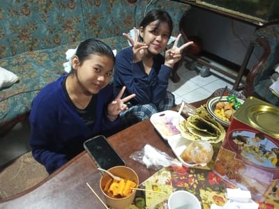 This image depicts two young females seated at a wooden table laden with various food items, inside a residence in Pandaria, India. The female on the left wears a dark blue cardigan over a black top, with her dark hair tied back with a white accessory; she smiles at the camera, forming a peace sign with her left hand. The female on the right wears a dark blue top and plaid trousers; she also smiles at the camera, making peace signs with both hands.

The wooden table is partially covered by a floral-patterned cloth. Visible items on the table include a black smartphone, a light brown paper cup or bowl containing orange skewered food, a white take-out container with purple and white food, a plastic bag containing food, a green-tinted flatbread, a white bowl of assorted fried snacks accompanied by small green packets, a red and gold-lidded tin depicting an illustration of people, and a white paper cup.

The background reveals a floral-patterned sofa (blue/green with yellow/brown accents) behind the subjects. To the right, an ornate wooden furniture piece, a white tiled floor, a grey dumbbell, and an orange basketball are visible. A glass tank or aquarium is partially visible in the upper right background. The scene is illuminated by interior lighting.
