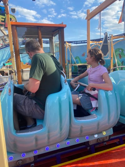 The image shows a man and a young girl on a amusement park ride in Baunatal, Germany. The ride appears to be a spinning teacup-style attraction, with the two passengers seated in a teal-colored car. The man, wearing a green t-shirt and grey shorts, is in the front seat and appears to be steering. The girl, in a pink and white striped shirt and denim skirt, is in the back seat, looking to her left and smiling. The background features a blue sky with white clouds, a wooden structure with orange trim, and a colorful mural with cartoon dinosaurs. The ride itself has blue lights on its base.