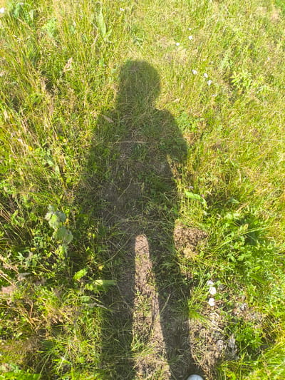 The image shows a shadow of a person cast on a patch of grass. The shadow is dark and elongated, suggesting the sun is low in the sky. The grass is green and appears to be growing in a somewhat uneven terrain, with patches of soil visible. There are also some small white flowers scattered among the grass. The lighting suggests it is daytime. The scene is located in Anastasivka, Ukraine.