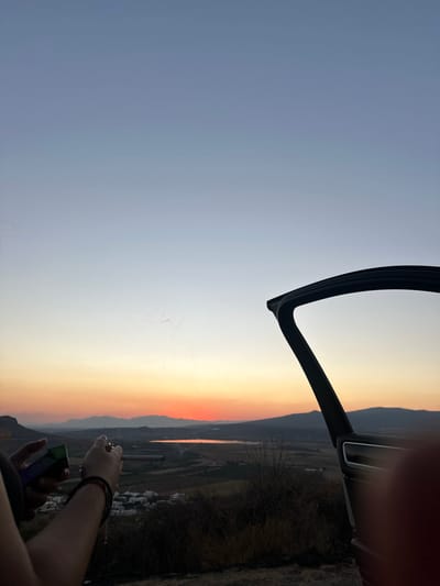A sunset view is framed by a car door's outline in the upper left. The sky transitions from blue to orange. A landscape is visible with a hint of mountains and a river, potentially with reflections of the sunset. A person's hand holding a phone is visible in the bottom right corner, with another hand visible slightly above it. The scene is likely taken from inside a car.
