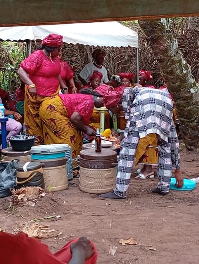 The image shows a group of people gathered outdoors in Umundugba, Nigeria, likely for a celebration or event. Several women wearing matching red tops, yellow skirts, and headwraps are congregated around food preparation. One woman, clad in a checkered outfit, bends over a table, possibly assisting with serving. Food containers and buckets are clustered on the ground, while others sit on a table and the containers. A partially visible white tent or canopy provides some cover. A tree trunk stands to the side. The ground appears to be dirt.