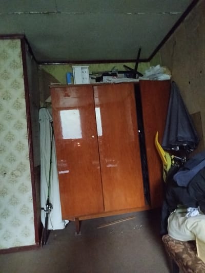 The image depicts a cluttered interior space, likely a room within a residential building in the Дзешчанскі сельскі Савет, Belarus. The focal point is a large, wooden wardrobe with two doors, exhibiting a reddish-brown finish. To the left of the wardrobe, a fishing rod is propped up against a white sheet or garment.
Above the wardrobe, on a shelf or surface, are various items including a white plastic container, possibly a small cabinet or dispenser, and other indistinct objects and papers. Part of a dark-colored umbrella and a yellow object are visible to the right of the wardrobe, partially obscuring other items like clothing.
The walls are covered in a patterned wallpaper on the left, transitioning to a less distinct, possibly damaged or discolored wall on the right. The ceiling appears to be textured and shows signs of wear, with a crack visible. The floor is a dark, nondescript surface with scattered debris. The lighting suggests an indoor environment, possibly with low natural light. The overall impression is one of disarray and storage.