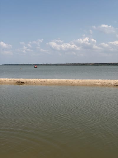 The image depicts a serene water scene, likely a bay or river, in Pavlo-Ochakovo, Russia. In the foreground, a narrow strip of sandy land juts out into the water, creating a visual division. The water itself is a muted green-brown, with subtle ripples indicating a gentle current. In the middle ground, on the surface of the water, a small red and white vessel is visible, possibly a boat or a floating structure. Further in the distance, a faint line of land stretches across the horizon, with some vegetation and possibly buildings dotting the landscape. The sky above is a pale blue, with scattered white, puffy clouds. The overall atmosphere is peaceful and natural, suggesting a calm day in a coastal or riverside environment.