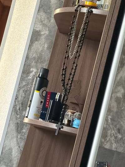 This is a close-up, high-angle shot of a wooden shelf containing various items. The shelf is mounted on a textured gray wall.
On the shelf, from left to right, are:
* A black aerosol can with a yellow and white label. The label has text including "FOR" and "INVISIBLE" and a circular graphic with "48".
* A blue and red box with the word "PRADA" printed in red.
* A black cologne bottle with a silver cap and a small silver nozzle.
* A string of dark beads, possibly a rosary, with silver chain links interwoven. The beads are predominantly black.
* A small silver cross charm is placed on the shelf near the cologne bottle.
* Two small glass jars are visible behind the cologne bottle. One has a cork stopper and contains a brown object. The other has a clear stopper and contains a small blue figurine, possibly a cat.
* A faint silhouette of a person's profile is carved into the wooden shelf backing behind the rosary.
* A small framed photograph is visible at the top of the shelf behind the rosary.
The foreground of the image is dominated by the shelf and its contents. The background consists of the textured wall and a portion of a white, vertical element, possibly a doorframe or a mirror. The lighting suggests an indoor environment. There are no people present in the immediate scene. The city of Avdarma, Moldova is not visually identifiable in this image.
