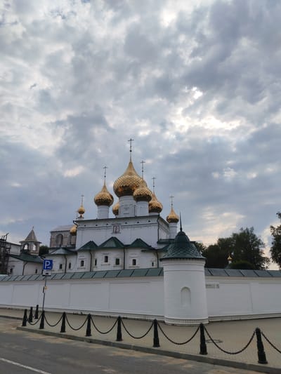 The image captures a view of the Assumption Cathedral in the city of Kostroma, Russia, not Vasil'ki. The cathedral, adorned with multiple golden domes topped with crosses, stands prominently against a cloudy sky. It is surrounded by a white brick wall, with a small, round, white tower integrated into the wall on the right side of the frame. A black chain-link fence with black bollards runs along the edge of a paved area in front of the wall. A blue parking sign is visible on a pole near the wall. The foreground shows a gray paved surface, possibly a road or parking lot, with white markings. Lush green trees are visible in the background to the right of the cathedral. The overall scene is one of architectural grandeur under a dramatic sky.