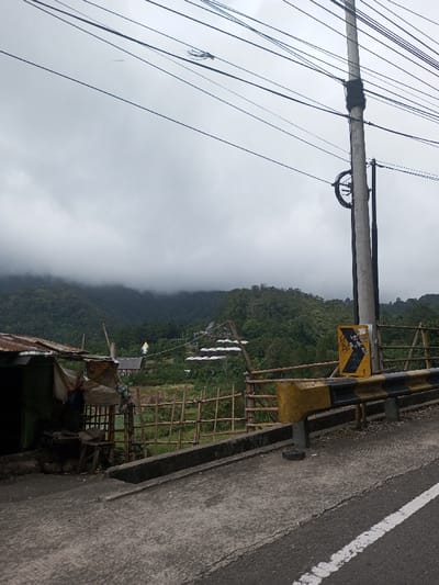 The image displays a scene in Saing Ta Mawt Lay, Myanmar, under an overcast sky. In the foreground, a paved road with a white demarcation line transitions to a concrete shoulder. A yellow and black striped guardrail runs along the right edge, behind which a grey utility pole stands. Numerous black electrical wires are strung across the upper portion of the frame, attached to the pole which also features a bundled section of material and a circular component. A yellow directional arrow sign, marked with black graffiti, is mounted to a post next to the guardrail. On the left, a structure with a rusty corrugated metal roof and tarpaulin walls is visible, alongside a wooden fence and a small wooden stool. A bamboo fence separates the immediate foreground from a vegetated area. In the midground, several white-roofed buildings are visible on a hillside, and a golden, tiered structure, likely a religious monument, stands out among the trees. The background consists of densely forested mountains, their upper sections obscured by thick, low-hanging mist or clouds. No individuals are present in the scene.
