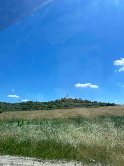 The image depicts a landscape on a clear, sunny day. The sky is a vibrant blue with scattered white cumulus clouds, suggesting fair weather. A large portion of the frame is occupied by the sky, with a notable reflection of what appears to be a vehicle's interior on the left side of the image.
The ground level shows a transition from a field of dry, light brown grass to a more lush, green, and taller grass on the right. Beyond the grassy areas, a line of trees and dense vegetation creates a natural barrier. In the distance, several tall, lattice-like electrical towers are visible, indicating the presence of power lines.
The scene appears to be a rural or semi-rural environment, possibly along a roadside given the perspective and the suggestion of a vehicle. The time of day is likely midday or early afternoon due to the bright sunlight and clear sky. There are no people or specific objects of interest, other than the natural landscape and the man-made electrical infrastructure. The overall mood is peaceful and serene, with no indication of any particular activity or event. No text is visible in the image. The provided location context is Tatarka, Russia.