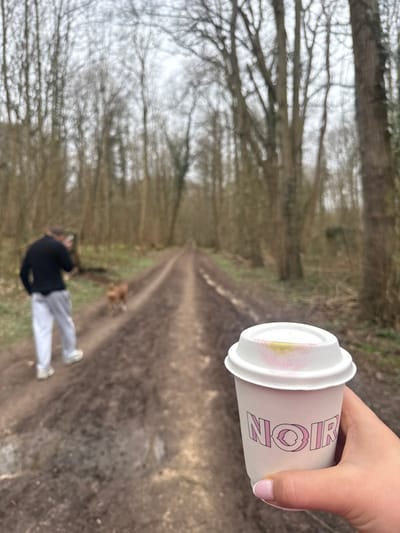 A dirt path stretches into the distance, flanked by bare trees under a cloudy sky. A blurry figure stands on the path, facing away. A hand holds a "NOIR" coffee cup with visible lipstick marks on the lid. The scene suggests an outdoor setting, possibly a forest trail. The cup's placement implies a focus on the beverage against the backdrop.