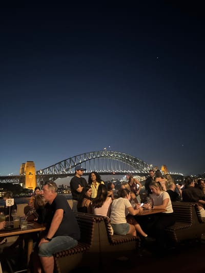 The image shows a nighttime view of the Sydney Harbour Bridge and part of the city skyline. A group of people are sitting at tables, likely at a bar or restaurant, enjoying the view. Many are looking at or interacting with their phones. Some are seated on benches. The sky is dark blue, and stars are visible. The bridge is illuminated, with lights outlining its structure. In the foreground, there are tables with glasses and other objects. One table has the number "23" printed on it.