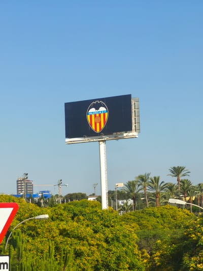 A wide shot of a clear blue sky above a billboard displaying the Valencia C.F. crest, supported by a tall white pole. The billboard is situated in a park-like setting with lush green trees and vegetation, some bearing yellow flowers, in the foreground and mid-ground. In the background, a tall, modern building with a dark facade stands to the left, alongside construction cranes and other smaller buildings. Palm trees are visible to the right of the billboard. A red and white triangular traffic sign is partially visible in the bottom left corner, along with a black sign with the letter "m". The scene appears to be outdoors during the day, likely in a city environment, possibly Paterna, Spain, given the presence of the Valencia C.F. crest.