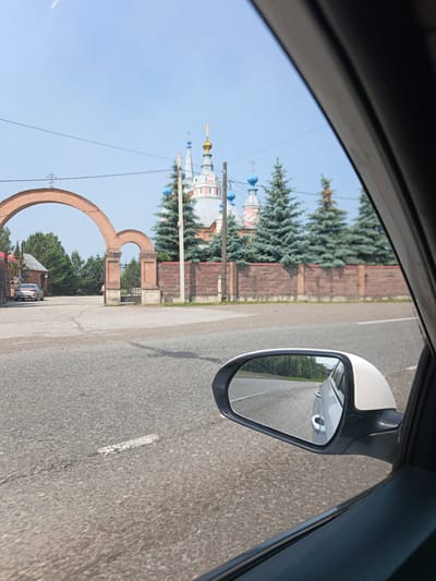 An eye-level, medium shot captures a view from inside a car in Bezrukova, Russia. The foreground features the car's side mirror reflecting the road and trees, with a sliver of the car's interior visible on the right. The middle ground displays an arched brick gateway leading into a courtyard. Beyond the gateway, a church with blue and gold domes and a red and white facade is visible, surrounded by evergreen trees and a brick wall. A silver car is parked to the left of the gateway, and a person is walking near the entrance. The background consists of a clear blue sky with a faint wire stretched across it. The road in the foreground is asphalt with a white line.