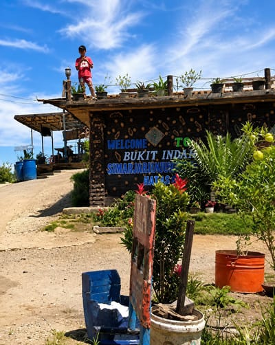 A young boy wearing a red t-shirt and shorts stands on the roof of a building. The building has a dark wooden facade decorated with circular patterns and a sign that reads "WELCOME BUKIT INDAH SIMARJARJUNJUN HATAS". Potted plants are arranged along the edge of the roof. The building is situated in an outdoor environment with a dirt path leading up to it. In the foreground, there is a blue chair, a weathered sign, a potted plant with red flowers, and an orange bucket. The sky above is blue with scattered clouds. The location is likely the city of Parik Sabungan, Indonesia, based on the sign.