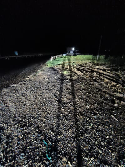 This is a nighttime photograph taken at an outdoor location, possibly in Thôn 9, Vietnam, given the context. The scene is dimly lit, with a light source from an unspecified direction casting long shadows across a gravelly, uneven surface. In the background, there appears to be a structure with lights on, suggesting a building or dwelling. A fence or barrier runs along the left side of the image, separating the road or path from the darker area beyond. The ground is covered in small stones and dirt, with some sparse vegetation visible. The overall impression is one of an undeveloped or rural area at night. There are no visible people in the frame, but the long shadows imply the presence of at least one person or object outside the camera's view. The image has a gritty, low-light aesthetic.