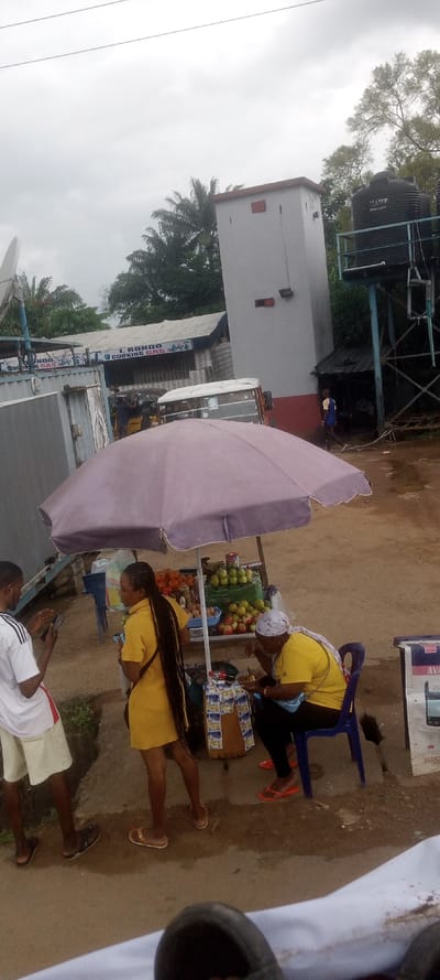 This image depicts a market scene in Ezinifite, Nigeria, under an overcast sky. The foreground is partially obscured by a dark object.
In the midground, a purple market umbrella provides shade over a fruit and produce stand. The stand displays various items including oranges, green apples, red apples, and eggs. Below the stand, packets of goods are hanging. Three individuals are gathered around this stand:
1. A male in a white short-sleeved shirt with black shoulder stripes and beige shorts stands on the left, looking at a mobile phone.
2. A female in a yellow short-sleeved dress with long dark braids stands facing the fruit display.
3. A second female, wearing a yellow short-sleeved shirt, dark pants, and a white headscarf, is seated on a blue plastic chair to the right of the stand, eating from a bowl with a spoon. She wears orange flip-flops and has a light blue bag slung over her shoulder.
In the background, multiple structures are visible. To the left, a grey shipping container-like structure and a satellite dish are present. Adjacent to this is a building with a corrugated metal roof and signage that reads "J. RONDO COOKING GAS." A light-colored tricycle-like vehicle is partially visible behind the market umbrella. To the right, a tall, white-painted utility building or tower features a red rectangular vent and two light fixtures. Next to this tower, a metal framework supports two large black water tanks, one labeled "KAPT." A person wearing a blue and yellow shirt is walking in the distance near these structures. The ground is a dirt or gravel road with some damp patches. Overhead, power lines traverse the scene. Lush green trees, including palm trees, are visible throughout the background.