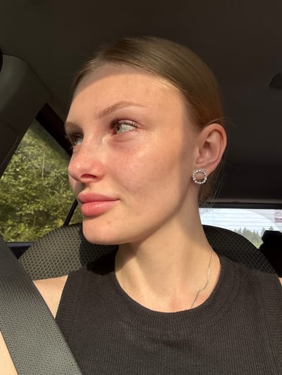 A young woman with light skin, blonde hair pulled back, and green eyes is pictured from the shoulders up, seated in the passenger seat of a car. She is looking to her left, away from the camera, with her head slightly tilted. Her lips are slightly parted, and she appears to be in profile. She is wearing a black sleeveless top and a thin silver chain necklace. A circular earring with small stones is visible on her right ear. A gray seatbelt is diagonally across her chest. Sunlight streams in from the left, illuminating her face and creating a shadow on the right side of her head. Outside the car window, lush green trees are visible. The interior of the car is dark, with the textured seat visible behind her. The image's lighting and the presence of trees suggest an outdoor setting, possibly a scenic drive. Without further information, it is not possible to definitively determine if this image was taken in Zarechnaya, Russia, as the visual elements are not specific to that location.