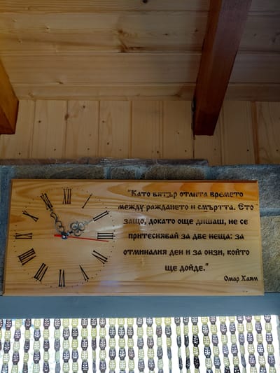 The image shows a wooden clock face with Roman numerals and text etched into it, hung on a stone wall with a wooden ceiling above. The text is in Bulgarian and appears to be a quote. The clock hands are visible, with a red second hand. Below the clock, there is a beaded curtain. The environment suggests an indoor or covered outdoor space, possibly a home or a rustic establishment. The location is specified as Dryanovets, Bulgaria.