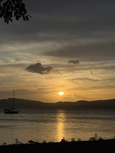 A sailboat is anchored in the calm waters of Lake Nicaragua, near Altagracia, Nicaragua, as the sun sets over the horizon. The sky is filled with dramatic clouds, tinged with shades of yellow, orange, and gray, creating a striking contrast with the dark silhouettes of the distant hills. The water reflects the vibrant colors of the sunset, with a shimmering golden path leading from the sun to the viewer. The foreground is a dark silhouette of the shoreline, with sparse vegetation and a few rocks visible. The overall mood is serene and peaceful, capturing the beauty of a tropical sunset.