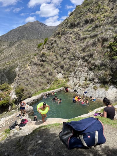 This image captures a natural swimming pool nestled within a mountainous, arid landscape near Mucurubá, Venezuela. The pool, constructed with stone walls, is filled with clear, green water and surrounded by rocky terrain covered in dry grass and sparse vegetation. A group of people, including adults and children, are enjoying the water. Some are swimming, while others are sitting or standing on the edge of the pool. A young boy holds a bright yellow inflatable ring. On the bank to the left, several pieces of clothing are laid out, along with a blue duffel bag in the foreground. The background features a steep, rocky mountain slope under a partly cloudy blue sky.