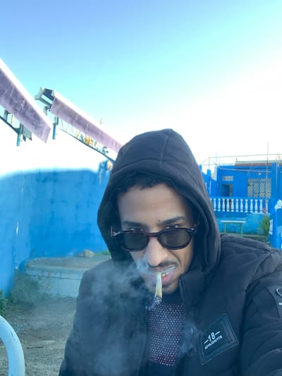 A person with sunglasses and a hood smokes a cigarette, exhaling smoke into the air. They wear a dark-colored jacket. In the background, there's a blue building with white railings and a clear blue sky. A structure with lavender awnings is also visible. The person appears to be outdoors during daylight.