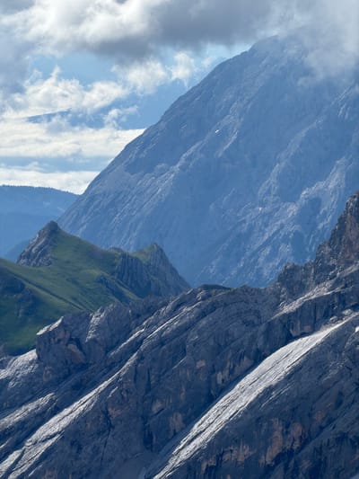 This is a photograph of a mountain landscape. The foreground shows steep, rocky slopes with some patches of scree and sparse vegetation. Behind these, a grassy, green mountain slope rises sharply, culminating in jagged peaks. Further in the background, a massive, imposing mountain dominates the scene, its rocky face deeply furrowed and textured, with some areas shrouded in clouds. The sky above is partly cloudy, with dramatic formations of white and grey clouds against a blue sky. The image conveys a sense of rugged natural beauty and the grandeur of alpine scenery. The location is the area of Wildermieming, Austria, as indicated by the provided information, and the image captures a view of the prominent mountain ranges in this region.