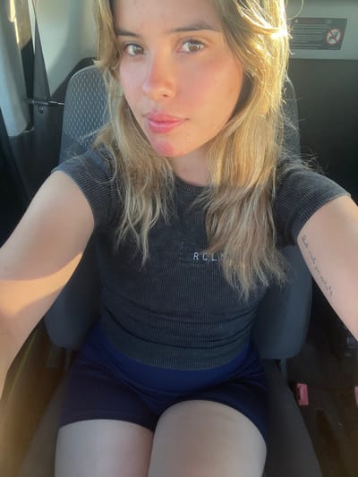 A female individual with long blonde hair and light skin is depicted seated inside a vehicle, likely a car or bus, taking a self-portrait. She is wearing a dark grey ribbed short-sleeved top with "RCLIN" and partially "PAUSA COLOMBIA" visible on the chest, and dark blue athletic shorts. Reddish blotches are present on her chin and nose. A script tattoo is visible on her left forearm. The interior features dark grey fabric seats, with strong sunlight entering from the left side of the frame, illuminating the subject's hair and right arm. A white decal featuring a red circle with a diagonal line over an image is visible in the upper right background. The specific location within Santa Rosa, Argentina, cannot be determined from the visual information provided.
