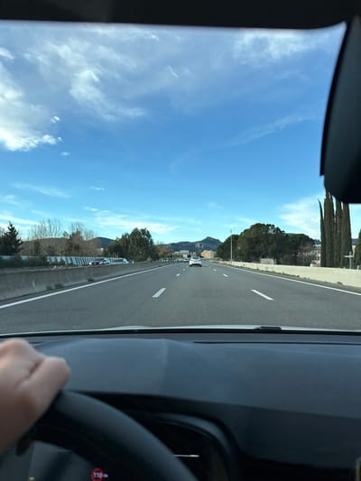 The view from inside a car moving on a highway. The clear blue sky dotted with white clouds is visible through the front windshield. Mountain peaks are seen in the distance beyond the trees lining the road. Cars are ahead on the multi-lane highway. A steering wheel and part of the dashboard are visible on the right side of the frame.