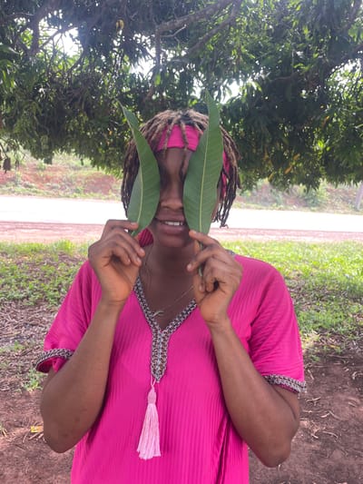 The image displays a person outdoors, captured in a playful moment under a lush tree canopy, likely in Ogbe, Nigeria. The individual, with medium-dark skin and dreadlocks, is wearing a vibrant hot pink tunic or dress. This garment features an elaborately embroidered neckline with silver-toned chain-link details, pink stitching, and a decorative pink tassel hanging from the front. They also sport a pink headscarf.
The central activity involves the person holding two large, green leaves – reminiscent of mango leaves – one over each eye, partially obscuring their face. A clear, happy smile is visible beneath the leaves, conveying a joyful or mischievous mood. Their hands, with well-manicured nails, are visible holding the leaves. Around their neck, a delicate silver-colored necklace with a distinctive pendant can be seen.
The setting is an unpaved, natural outdoor environment. Densely packed green foliage dominates the background and upper sections of the image, suggesting the person is standing under a large tree. A dirt path or road is visible further back, separating areas of green grass and scattered dry leaves on the ground. The lighting indicates it is daytime, with sunlight filtering through the leaves, creating soft highlights and shadows. No visible text is present in the image.