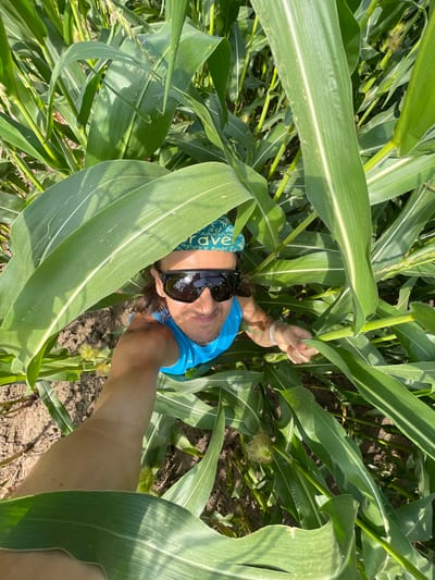 A man is taking a selfie in a cornfield. He is wearing a blue tank top and sunglasses, and a bandana on his head. The corn stalks are tall and green, and the ground is covered in dirt. The man is smiling and looking at the camera. The image was taken in Perevoznoe, Russia.