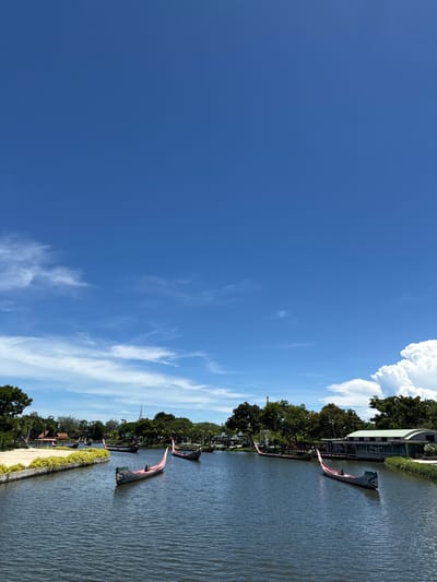 The image depicts a waterway in Bang Pu Subdistrict Municipality, Thailand, under a bright blue sky with scattered clouds. The water surface is rippled and calm. Along the waterway, there are trees, vegetation, and architecture resembling buildings. Gondola-like boats, decorated with red accents, are docked along the edge.