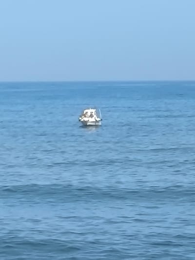 A white motorboat is anchored in the vast, blue ocean. The boat appears small against the expansive sea, with a cabin structure visible. Several figures are present on the boat. The water is calm, with gentle ripples across the surface. The sky is a clear, pale blue, suggesting a bright, possibly mid-morning or afternoon, with good weather. The scene conveys a sense of peaceful solitude at sea. The location is identified as La Gaspara, Spain. No text is visible in the image.