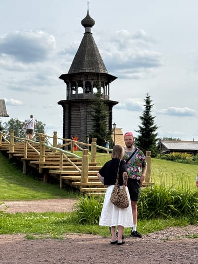 In Nevskii Parkleskhoz, Russia, a group of people are gathered near a wooden bell tower and a wooden staircase. A man wearing a pink cap and a colorful floral shirt stands next to a woman in a black top and white skirt, who is carrying a leopard-print bag. Further up the wooden staircase, a man in a white shirt and shorts walks away from the camera. In the background, there are trees, buildings with wooden roofs, and a cloudy sky. The ground is covered with gravel and grass.