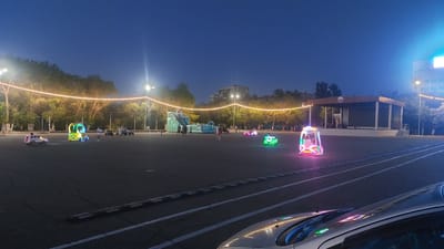 In the city of Priozersk, Kazakhstan, a nighttime scene unfolds in what appears to be an outdoor amusement or recreational area. The sky is a deep, clear blue, transitioning to dark, indicating dusk or early evening. A string of warm, yellow lights hangs across the upper portion of the frame, illuminating the tops of trees and creating a festive atmosphere.
In the foreground, the asphalt ground is marked with white lines, possibly designating a track or parking area. A silver car is partially visible in the lower right corner, its reflective surface catching some of the ambient light.
Scattered across the open space are various amusement attractions. To the left, a white toy car with a driver is in motion. Further back, a brightly colored, multi-sided amusement ride with green and yellow lights is active, with at least one person visible inside. Nearby, a blue inflatable slide structure with a climbing wall and multiple slides is set up, and a few people are gathered around its base.
In the middle ground, other illuminated rides are present, including a green, car-shaped ride and a white and pink capsule-shaped ride, both occupied by individuals. A small stage or platform with a roof and columns stands in the background, with a few figures visible on or near it. The overall impression is of a lively outdoor space in Priozersk, Kazakhstan, after dark, filled with illuminated entertainment for visitors.