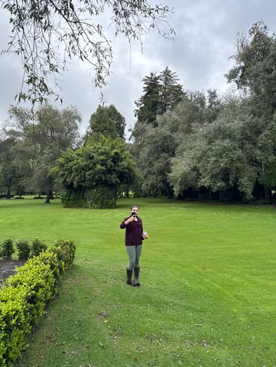 This image depicts a solitary adult female standing in a large, well-maintained grassy area under an overcast sky, located within a park or estate in Huasca de Ocampo, Mexico. The woman, positioned in the mid-right portion of the frame, wears a dark maroon long-sleeved top, light olive-green leggings, and dark tall rain boots. Her dark hair is pulled back. She holds a dark, rectangular object (likely a mobile phone or camera) with her right hand, raised to her face as if taking a self-portrait or video, while her left hand holds a white cylindrical object (possibly a cup or container).
In the immediate left foreground, a low, manicured green hedge lines a dark, undefined area. The expansive green lawn, featuring scattered small light-colored debris, extends towards a dense background of mature trees of varying species and foliage colors, including tall conifers and broadleaf trees, forming a natural border. A prominent, densely green, roughly mushroom-shaped tree or large bush, with some darker foliage or vines at its base, is situated in the mid-left background. The sky above is uniformly grey and cloudy. Bare branches from an overhead tree extend into the top portion of the frame, partially obscuring the sky.
