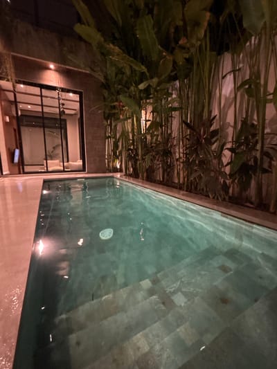 An eye-level, slightly angled photograph captures a swimming pool located in Tumbak Bayuh, Indonesia. The foreground features the pool's surface, reflecting ambient light and showing a slightly murky appearance. The pool's edge is constructed of what appears to be concrete.
To the left of the pool, lush tropical foliage hangs over a portion of the scene. Below the foliage, there's a building with large windows, offering a glimpse into an interior space. The building’s design appears modern. No people are visible in the image. The overall impression is of a tranquil, tropical setting.