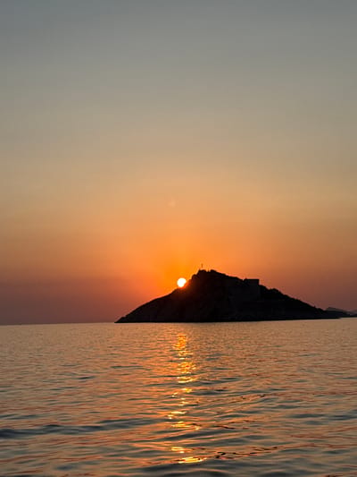 This is a vertical, eye-level, full-frame photograph taken at sunset in Mirišta, Montenegro. The image captures a serene seascape with a silhouetted island in the distance. The sun, a bright orange orb, is setting behind the island, casting a warm glow across the sky and water. The sky transitions from a soft gray at the top to a vibrant orange near the horizon. The water in the foreground reflects the sunset's colors, creating shimmering streaks of light on its surface. The island, with its rugged topography, is almost entirely in shadow, but a few structures are discernible on its peak. The overall mood is tranquil and picturesque.