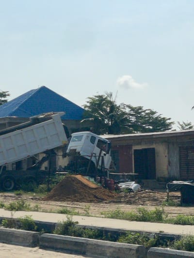 This image captures an outdoor scene in Igando Oloja, Nigeria, under a bright, clear sky with minimal cloud cover. In the mid-ground, a white dump truck is positioned with its empty cargo bed raised, indicating it has either just completed or is in the process of discharging material. Immediately adjacent to it on the right, a separate white truck cab sits tilted on a frame, suggesting it is either undergoing repair, assembly, or awaiting salvage. A prominent pile of reddish-brown sand or dirt is situated directly in front of these vehicles.

Behind the vehicles, two structures are visible. To the left, a building with a distinctive bright blue pyramidal roof is partially obscured. To the right, a single-story building with light-colored walls features multiple dark brown door or window openings. Various miscellaneous items, including white rectangular objects and red cylindrical containers, are scattered on the ground near the buildings and trucks. The foreground consists of a paved surface bordered by concrete curb barriers, beyond which lies a dirt area interspersed with sparse green vegetation. Additional green foliage, including palm trees, is visible in the background above and between the buildings. No individuals are present in the frame. The overall environment suggests a construction site or a vehicle repair/storage yard.