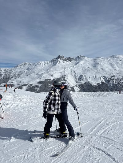 The image depicts a snow-covered mountain ski slope in Saint-Martin-de-Belleville, France. In the foreground, two individuals are positioned on ski equipment. The person on the left wears a black and white checkered jacket, black pants, a grey helmet, and dark goggles, standing on a snowboard. The person on the right wears a grey patterned jacket, dark pants, a dark helmet, and purple-tinted goggles, holding a ski pole in their right hand and standing on skis. Both individuals are leaning towards each other, with their faces largely obscured by helmets and goggles, appearing to be in a close embrace. The background features extensive snow-covered mountains, with visible peaks and scattered evergreen trees on lower slopes. The sky is partly cloudy. Numerous ski tracks are present on the snow. In the mid-ground to the left, several other skiers are visible further down the slope, along with a partial view of a ski lift structure, including an orange support pole.