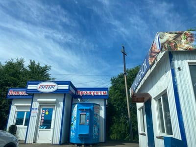 Two adjacent, small commercial kiosks, likely part of a roadside market in Buzuluk, Russia, are captured from a low-angle perspective under a bright blue sky with wispy clouds. The kiosk on the left has a sign with blue lettering that reads "ПИВЦ" followed by another word partially obscured. It features a white door with a window, a smaller window to the left, and a blue and white striped awning above. Below the awning, a sign displays operating hours: "РЕЖИМ РАБОТЫ" from 8:00-20:00, with a break from 13:00-14:00, and another entry of 17:45-18:00. A blue water vending machine is situated between the two kiosks, displaying images of water bottles and text, including "ВОДА ПИТЬЕВАЯ АРТЕЗИАНСКАЯ" and "24 ЧАСА". The kiosk on the right has a large banner above it displaying images of shawarma and other food items. Its side is made of corrugated white metal with blue trim, and it has two windows. A dark wooden awning protrudes from the right kiosk. In the background, lush green trees and a utility pole with wires are visible. A silver car is partially visible in the lower left corner.
